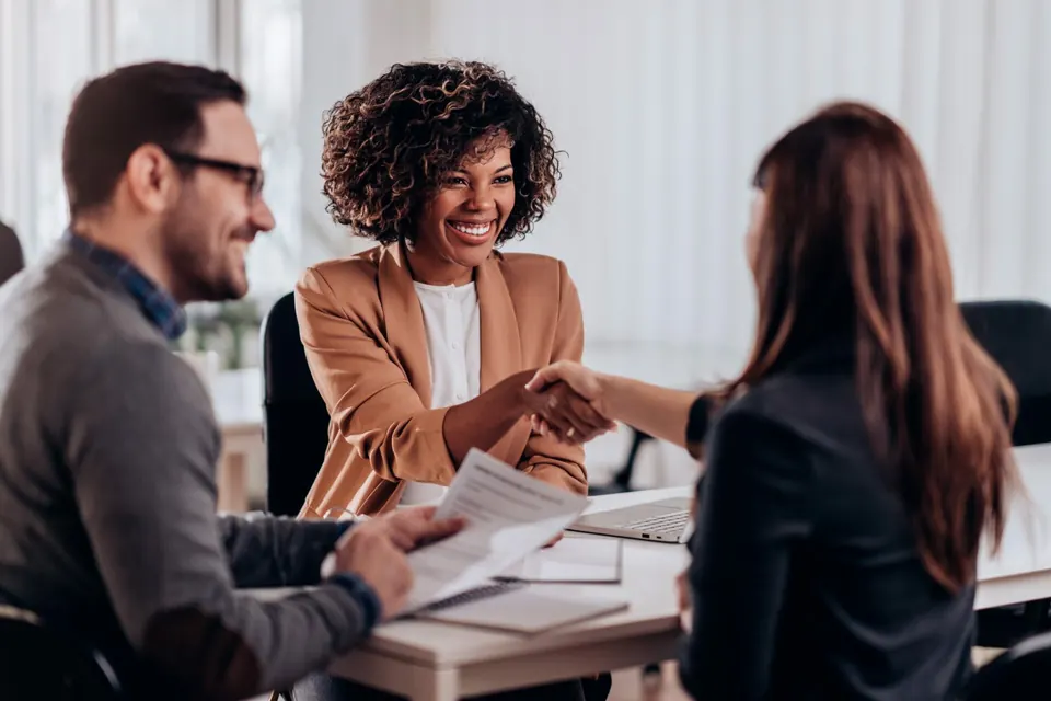 Três pessoas conversando e sorrindo em uma reunião; duas delas apertam as mãos